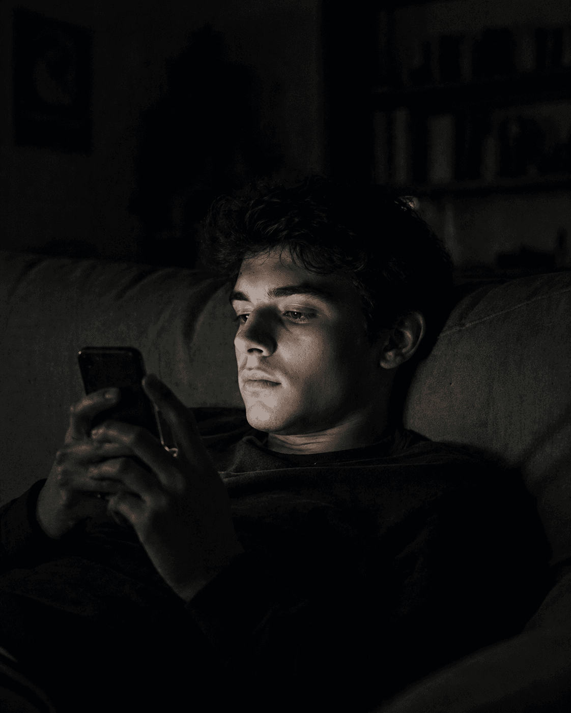 A young man in a dark room looking at a glowing phone screen, illustrating distraction and endless consumption